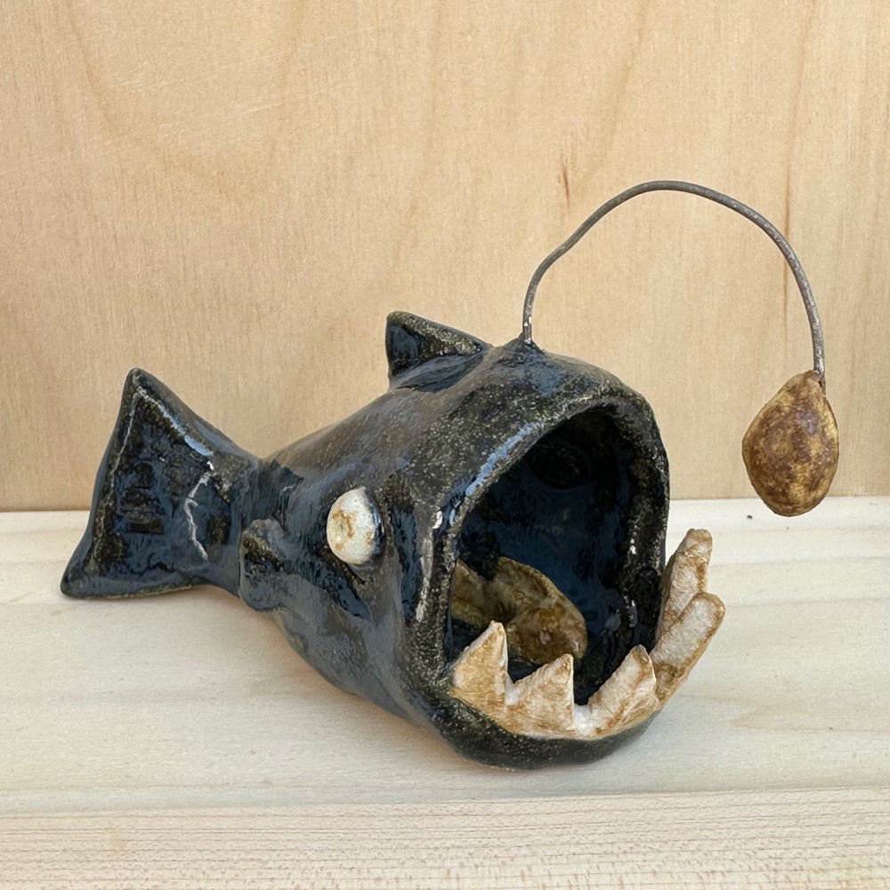 Hand built angler fish figure that features exaggerated large teeth, a dangling lantern light, and a small fishy inside it's gaping mouth